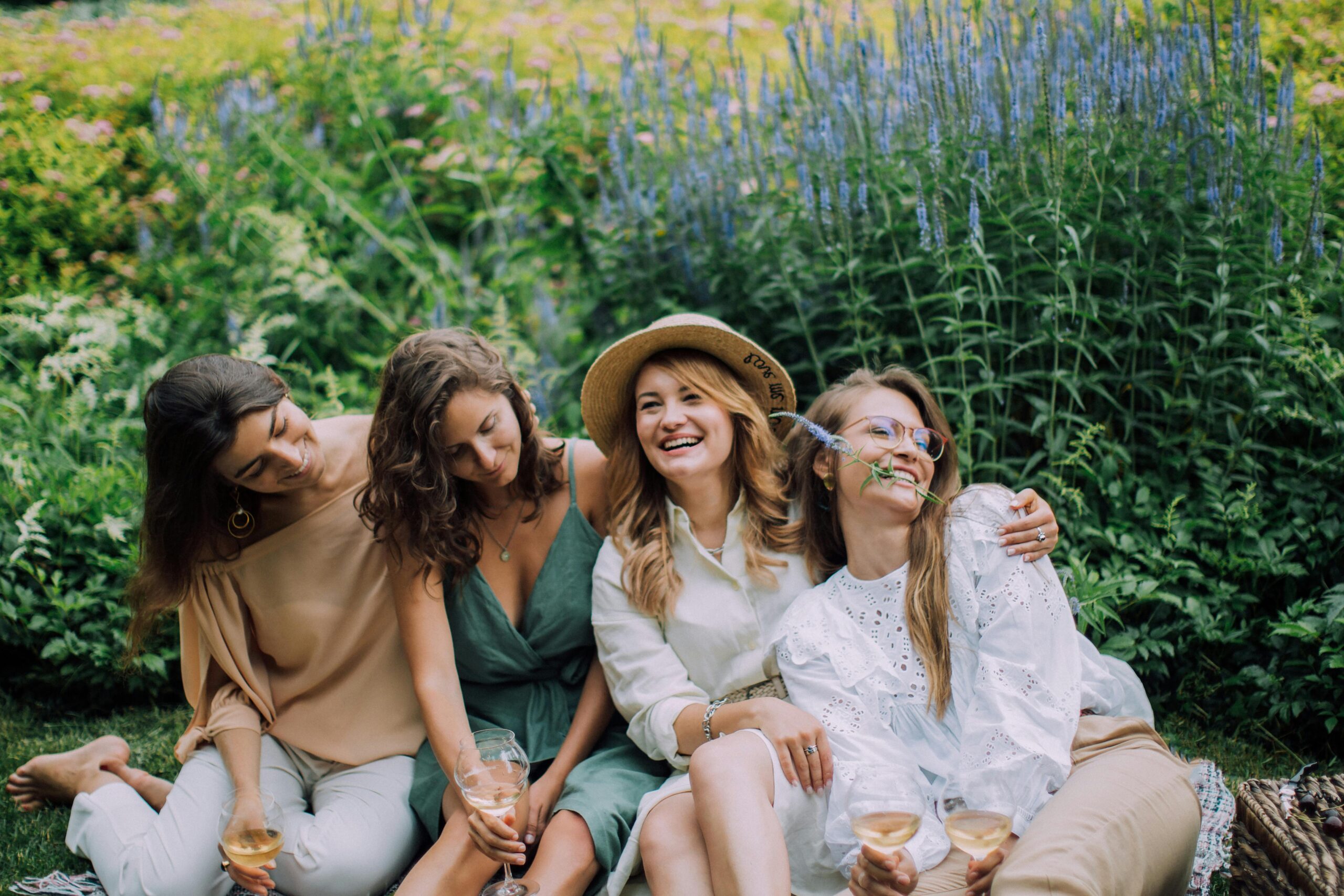 Four women enjoying a cheerful picnic with wine in a vibrant garden setting.